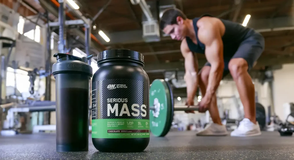 Optimum Nutrition Serious Mass protein powder tub and shaker bottle on a gym floor with a person lifting weights in the background.